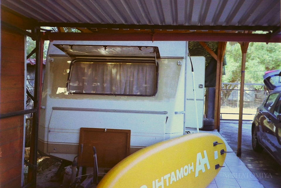 Small white camper trailer parked under a metal carport, with its window curtains visible and the awning partly extended. A yellow surfboard rests in front of the trailer, and a dark car with an open trunk is parked beside it. Trees and fencing appear in the background.